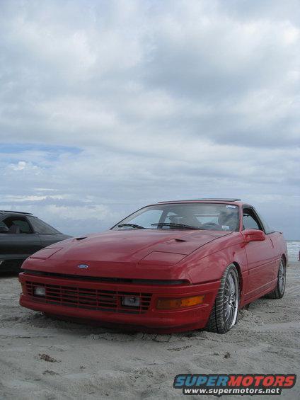 Red coupe with pop-up headlights on a beach, front 3/4 view. Another dark car is visible to the left