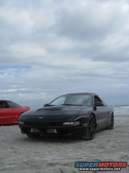 Black 1994 Ford Probe parked on a sandy beach with a red car in the background