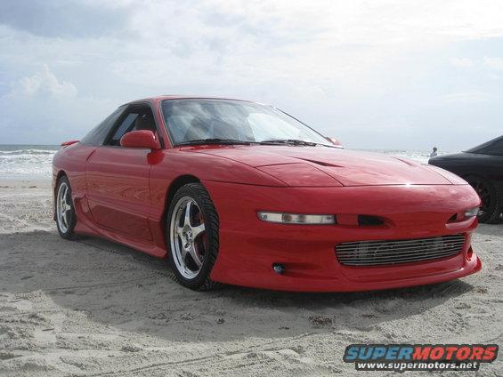 Red 1994 Ford Probe coupe parked on a sandy beach with the ocean in the background