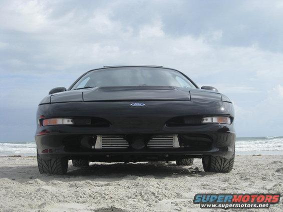 Front view of a black 1994 Ford Probe parked on a sandy beach with the ocean in the background