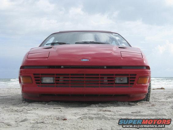 Front view of a red Ford Probe parked on a sandy beach with the ocean in the background
