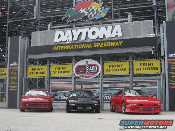 Three sports cars, two red and one black, parked in front of the Daytona International Speedway entrance