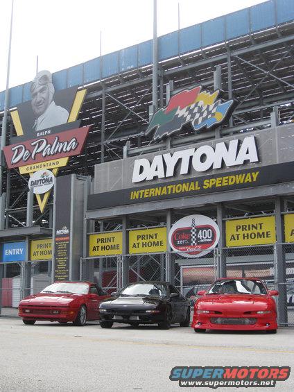 Three sports cars parked in front of Daytona International Speedway grandstands. Signs for Ralph De Palma and Coke 400 are visible