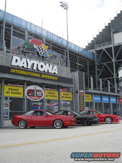 Three sports cars parked in front of Daytona International Speedway. The cars are red, black, and red
