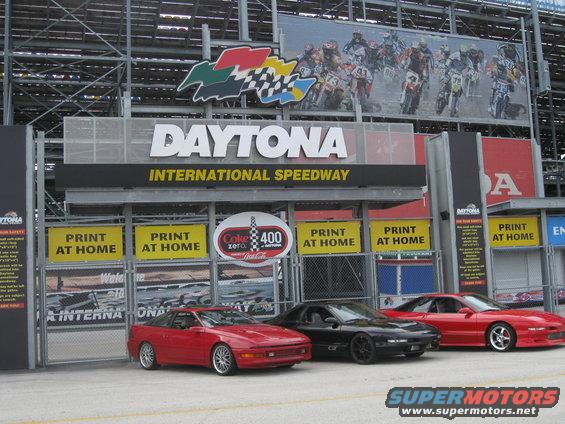Three sports cars, two red and one black, parked in front of the Daytona International Speedway entrance