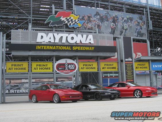 Three coupes, two red and one black, parked at Daytona International Speedway. The red car on the left has silver wheels