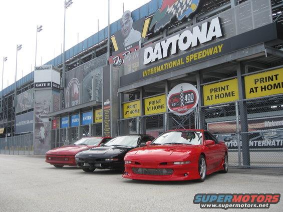 Three sports cars parked in front of Daytona International Speedway entrance. Red car in foreground
