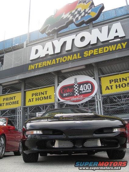 Black sports car parked in front of Daytona International Speedway sign. Another red car is visible to the left