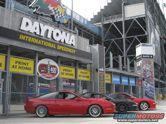 Three Ford Probe cars parked in front of Daytona International Speedway entrance. The lead car is red, followed by a black one, and another red one