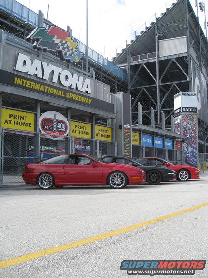 Three coupes parked in front of Daytona International Speedway entrance. The first car is red, the second is black, and the third is red