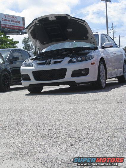 White Mazda 6 sedan with hood open, front view. Black car partially visible to the left