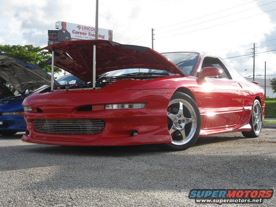 Red sports coupe with hood open, parked next to a blue car. Billboard in background