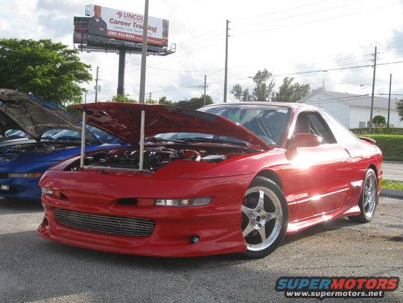 Red coupe with hood open, showing engine bay. Chrome five-spoke wheels. Another blue car with hood open behind it. Billboard in background