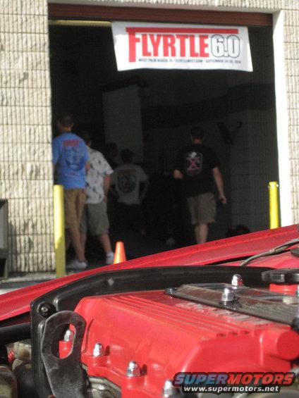 Red engine cover with chrome bolts and a metal bracket in the foreground. A banner reading "FLYRTLE 6.0" hangs above an open doorway