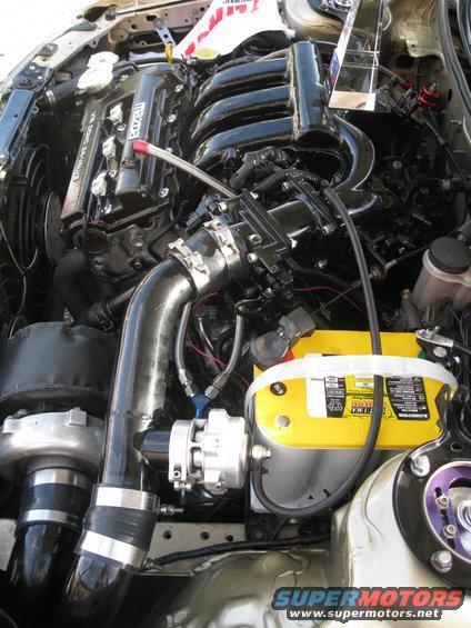 Engine bay with a turbocharger, black intake manifold, and yellow Optima battery. Some red and blue fittings are visible