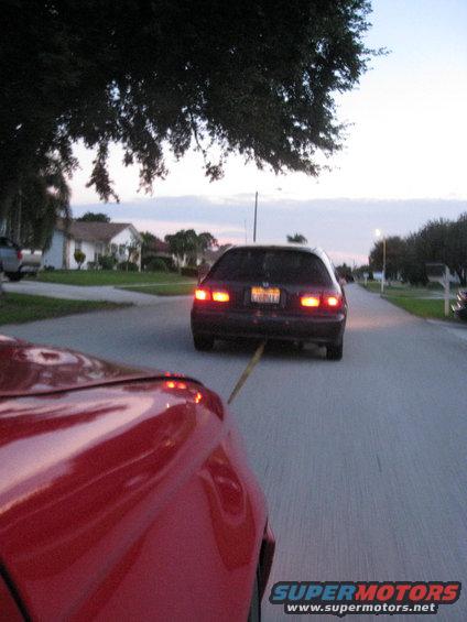Red car's front fender and hood in foreground, towing a dark hatchback with its taillights on