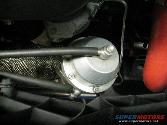 Silver turbocharger wastegate with black hoses and heat-wrapped exhaust pipe. Red silicone hose visible on the right. Text reads SUPERMOTORS
