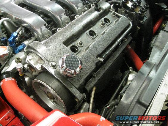 Engine bay showing a textured valve cover, chrome oil cap, red silicone hoses, and timing belt