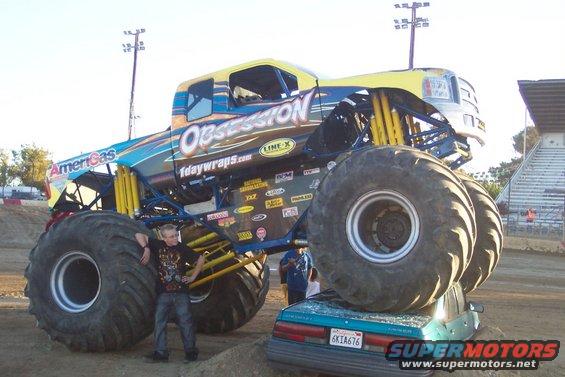 Monster truck named Obsession with large tires crushing a teal car. A person stands next to the front tire