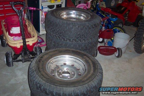 Stack of four tires with chrome rims in a garage, next to a red wagon and other items