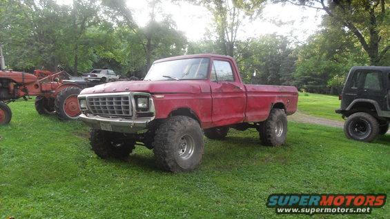 Red 1979 Ford F-150 pickup truck with large tires, parked on grass next to a tractor and a black Jeep