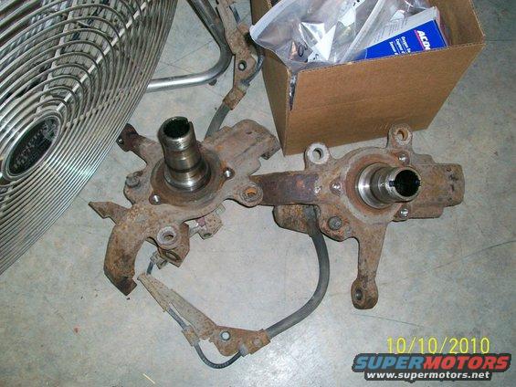 Two rusty vehicle steering knuckles with brake lines attached, next to a fan and a box of parts
