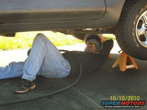 Man lying on the ground working under a vehicle supported by jack stands. Date stamp shows 10/10/2010
