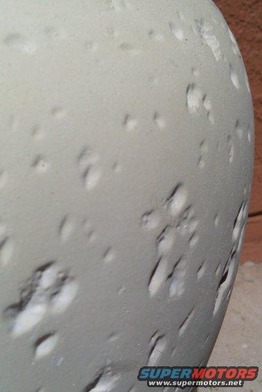 Close-up of a gray, textured surface with many small dents and imperfections. A watermark reads 'SUPERMOTORS.NET'