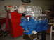 1965 Ford 427 - A blue V8 engine with a prominent supercharger is displayed in a workshop setting that appears to be a workshop.