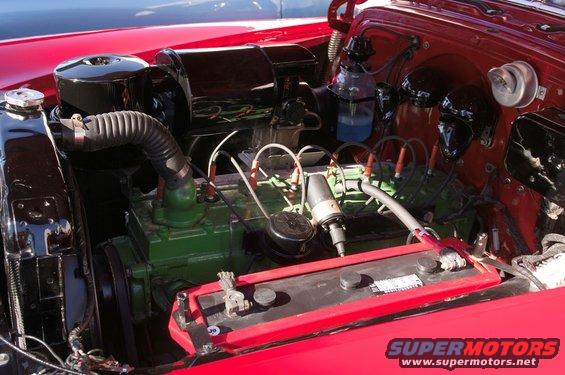 Engine bay of a classic red car, showing the green engine block, distributor, and battery