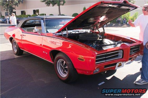 Red 1969 Pontiac GTO convertible with hood open, showing the engine. People are standing around it