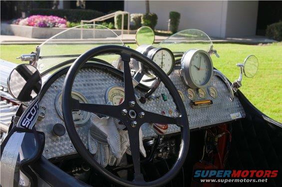 Vintage car dashboard with a four-spoke steering wheel, multiple gauges, and a clock. 'SCOT' is visible on a strap