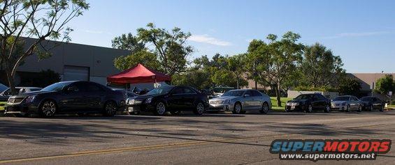 Several Cadillac sedans parked in a row, some black, one silver. A red tent is behind them