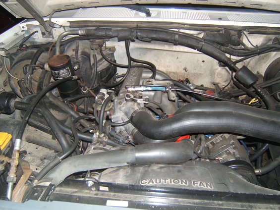 Engine bay with hoses, wires, and engine components visible. Fan shroud has 'CAUTION FAN' text