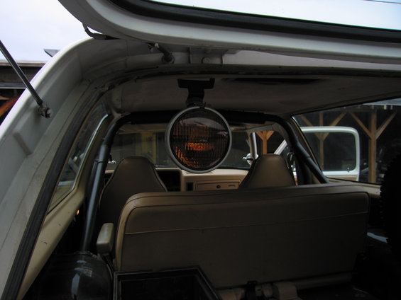 Interior view of a vehicle showing tan seats, a roll bar, and a mounted spotlight above the dashboard. Another vehicle is visible through the rear window
