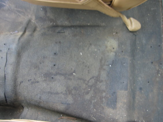 Car floor carpet with stains and some debris. A tan seat is visible at the top