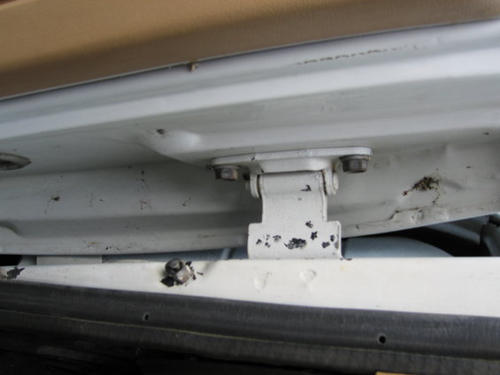 White door hinge with chipped paint, attached to a white vehicle frame. Visible bolts and interior trim
