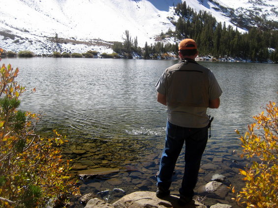 Man wearing a vest and orange cap fishes in a clear lake with rocky shore; snow-covered mountains and pine trees in background
