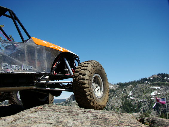 Off-road buggy with 'SLUT ME' text on the side, parked on rocks with mountains and an American flag in the background