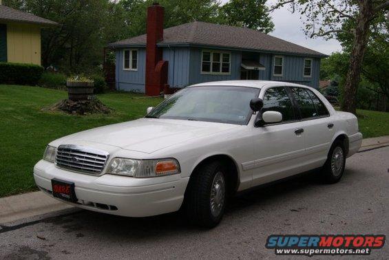 White sedan parked on a residential street. It has a police-style spotlight on the driver's side mirror