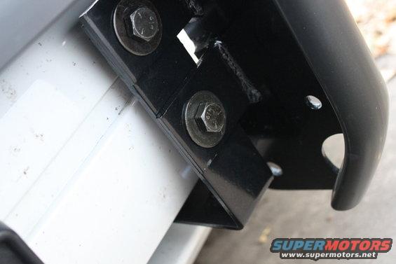 Close-up of a black metal bracket bolted to a white surface, part of a push bar assembly