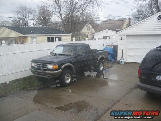 Black 1995 Ford Ranger pickup truck parked in a wet driveway next to a white fence and garage
