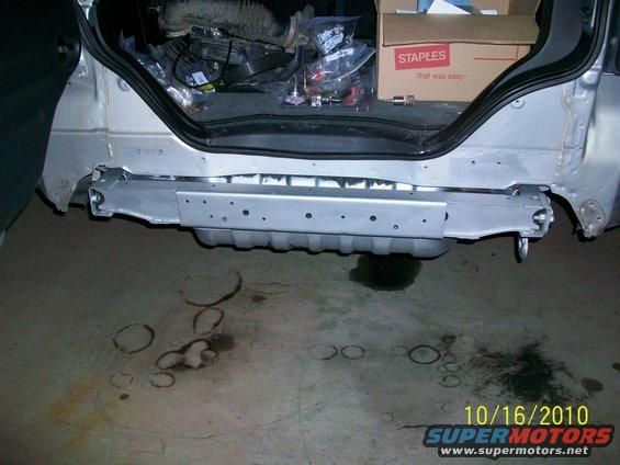 Silver vehicle's rear bumper assembly removed, showing structural beams and cargo area with parts and a Staples box