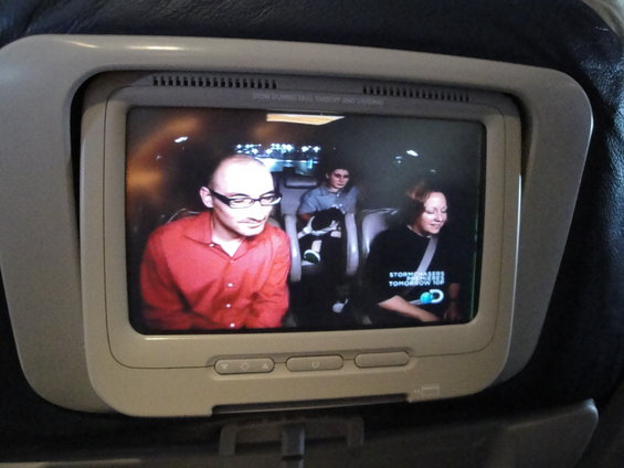 Airplane screen shows three people watching a video. One man wears a red shirt and glasses