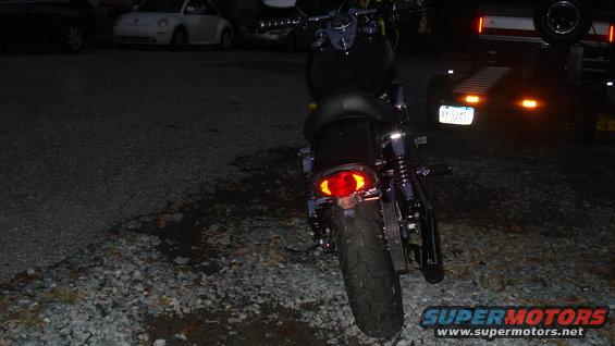 Motorcycle rear view, dark, gravel lot, trailer with lights in background