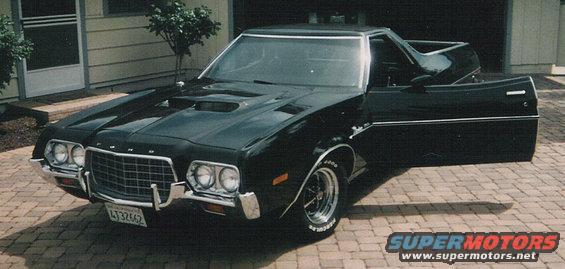 Black 1972 Ford Ranchero with open driver's door and hood scoop, parked on brick driveway