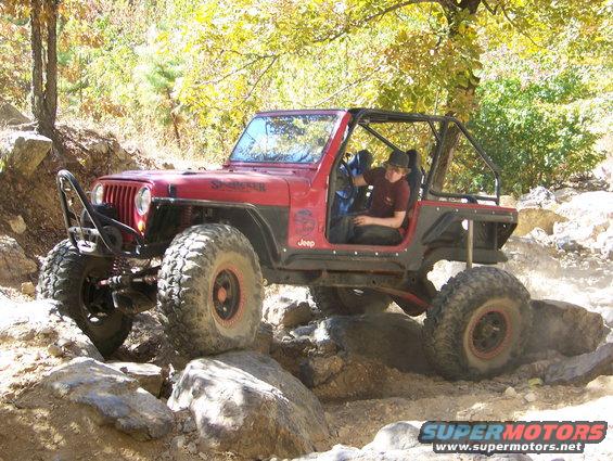 Red Jeep Wrangler with roll cage driving over rocks. Driver wearing a hat