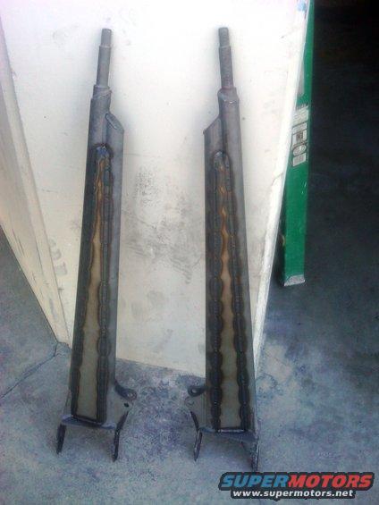 Two welded radius arms leaning against a wall. A green ladder is visible in the background