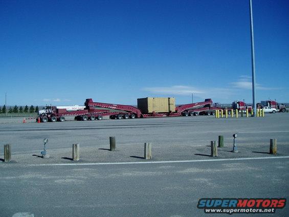 Maroon semi-trucks with long, multi-axle trailers carrying a large box under a clear blue sky