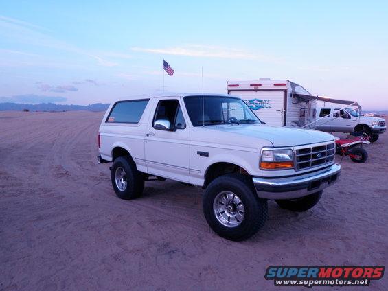 White Ford Bronco with aftermarket wheels parked on sand, 3q-front view. Trailer and truck in background